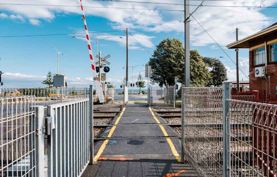 A pedestrian pathway leading across a railway crossing with metal safety gates, warning signs, and overhead power lines, set against a backdrop of trees, traffic signals, and a clear blue sky.